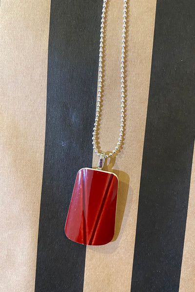 The Saved & remade necklace Porslin by BUTIK KUBIK features a silverplated chain with a shiny, trapezoid red pendant made from upcycled porcelain, shown against alternating black and tan vertical stripes.