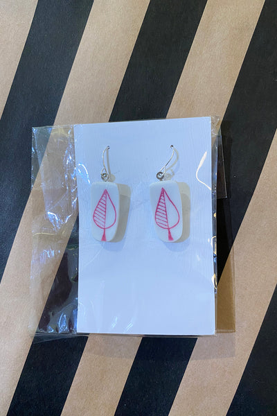 A pair of rectangular white Saved & remade earrings by BUTIK KUBIK, featuring pink leaf designs on återbrukat porslin, are shown on a white card in clear plastic packaging atop a black and tan striped surface.