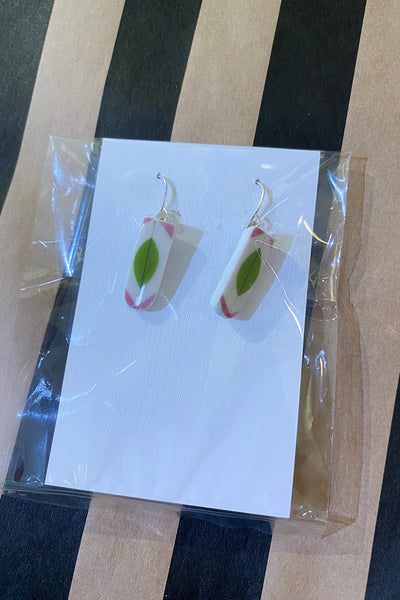 A pair of rectangular Saved & Remade earrings by BUTIK KUBIK, made from recycled porcelain with white, green leaf, and pink details. Presented on a white card in clear packaging, set on a striped brown and black surface.