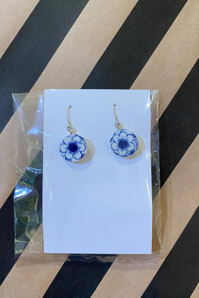 A pair of round Saved & Remade earrings by BUTIK KUBIK, crafted from recycled porcelain with blue and white floral designs, displayed on a white card in clear plastic against a black and tan striped background.