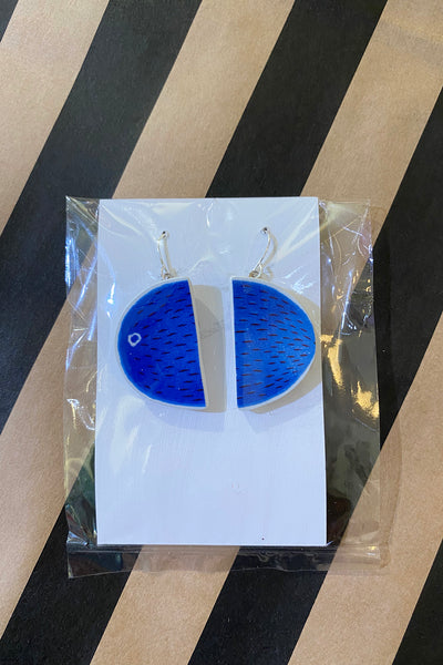 A pair of blue, semi-circular Saved & remade earrings by BUTIK KUBIK, crafted from återbrukat porslin with textured lines and a white dot, arranged in a circle on white card inside clear packaging against a striped background.