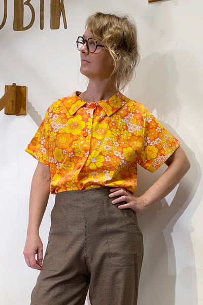 A woman with wavy blonde hair and glasses models the Kubik blouse vintage by BUTIK KUBIK—an oversized upcycled floral top in bright orange and yellow—paired with high-waisted brown pants, standing confidently against a white wall.