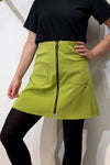 A person in a black short-sleeve top, black tights, and the BUTIK KUBIK Kubik zip skirt nr 1 Vintage green with front pockets and a center zipper stands confidently with one hand on their hip.