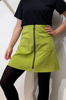A person in a black short-sleeve top, black tights, and the BUTIK KUBIK Kubik zip skirt nr 1 Vintage green with front pockets and a center zipper stands confidently with one hand on their hip.