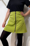 A person in a black short-sleeve top, black tights, and the BUTIK KUBIK Kubik zip skirt nr 1 Vintage green with front pockets and a center zipper stands confidently with one hand on their hip.