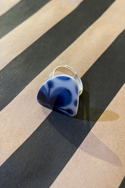 The Saved & remade ring porslin by BUTIK KUBIK features återbrukat Mon Amie blue and white ceramic, silverpläterad band, and rests on black and beige diagonal stripes—showcasing unique, sustainable style.