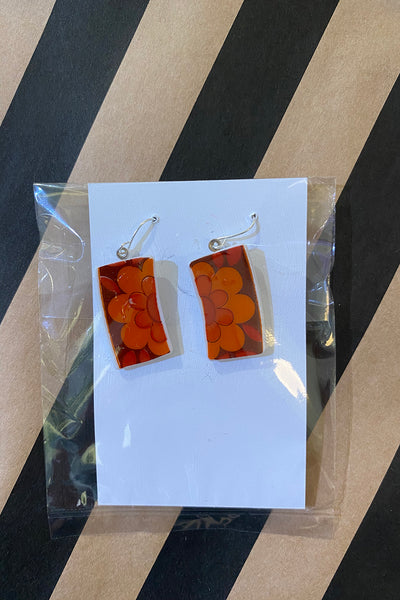 A pair of rectangular orange Saved & remade earrings by BUTIK KUBIK, featuring floral designs on upcycled porcelain, are shown on a white card wrapped in clear plastic atop a diagonal black and tan striped surface.