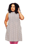 A curly-haired woman wears the Cissi och Selma dress Rut Petit Brun from BUTIK KUBIK with pink leggings, smiling and touching her hair while standing against a white background, one hand in her pocket.