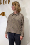 A person with blonde, wavy hair and glasses is wearing the BUTIK KUBIK Kubik blouse checkered with black pants, standing against a white wall with part of a wooden sign visible in the background.