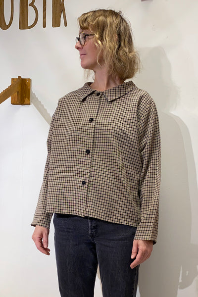 A person with blonde, wavy hair and glasses is wearing the BUTIK KUBIK Kubik blouse checkered with black pants, standing against a white wall with part of a wooden sign visible in the background.