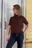A person with shoulder-length blonde hair and glasses stands indoors, wearing the BUTIK KUBIK Kubik t-shirt checked in a 60's inspired brown and black pattern, paired with dark jeans, looking to the side with one hand on their hip.