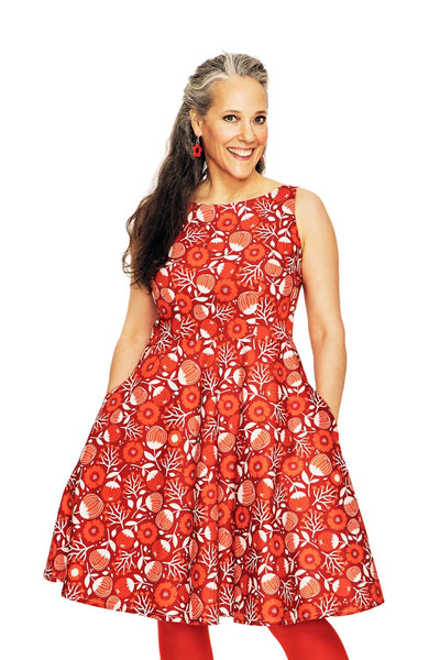 A smiling woman with long, dark hair streaked with gray wears the Cissi och Selma dress Saga Hassel Röd by BUTIK KUBIK, matched with red earrings and tights. She stands hands in pockets against a white background.