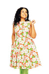 A woman with curly hair smiles in a Cissi och Selma dress Saga Humlesurr Korall from BUTIK KUBIK, featuring a bright orange, white, and green floral 50s-style pattern. She poses against a white background, holding her dress.