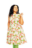 A woman with curly hair smiles in a Cissi och Selma dress Saga Humlesurr Korall from BUTIK KUBIK, featuring a bright orange, white, and green floral 50s-style pattern. She poses against a white background, holding her dress.