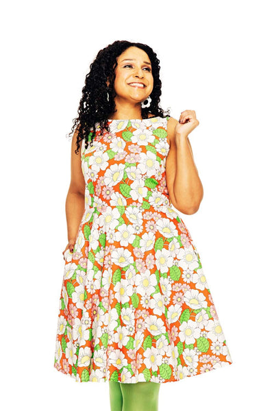 A woman with curly hair smiles in a Cissi och Selma dress Saga Humlesurr Korall from BUTIK KUBIK, featuring a bright orange, white, and green floral 50s-style pattern. She poses against a white background, holding her dress.