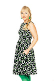 A woman with blonde hair in a side braid wears the "Cissi och Selma dress Saga Retroblom Svart" by BUTIK KUBIK, featuring a black base with white and green flowers. She smiles, one hand in her pocket, against a white background.