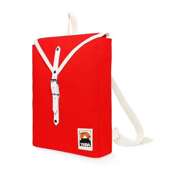 The BUTIK KUBIK YKRA Scout red is a small, bright red rectangular cotton canvas backpack with a white strap and buckle, a mountain logo patch with Ykra text on the front, and adjustable white shoulder straps—perfect for everyday city use.