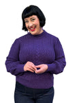 A smiling woman with short black hair wears the Cissi och Selma tröja Signe Lila by BUTIK KUBIK with ribbed cuffs and dark jeans, standing against a white background.