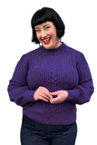 A smiling woman with short black hair wears the Cissi och Selma tröja Signe Lila by BUTIK KUBIK with ribbed cuffs and dark jeans, standing against a white background.