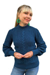 A smiling woman with blonde hair and bangs wears green earrings, light blue jeans, and the "Cissi och Selma tröja Signe Ocean" from BUTIK KUBIK, featuring a textured knit and ribbed cuffs. The background is white.