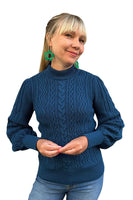 A smiling woman with blonde hair and bangs wears green earrings, light blue jeans, and the "Cissi och Selma tröja Signe Ocean" from BUTIK KUBIK, featuring a textured knit and ribbed cuffs. The background is white.
