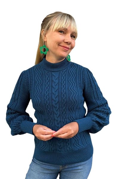 A smiling woman with blonde hair and bangs wears green earrings, light blue jeans, and the "Cissi och Selma tröja Signe Ocean" from BUTIK KUBIK, featuring a textured knit and ribbed cuffs. The background is white.