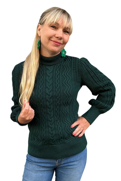 A smiling woman with long blonde hair wears the BUTIK KUBIK Cissi och Selma tröja Signe Skog, green earrings, and blue jeans, standing against a white background.