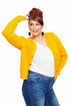 A smiling woman with brown hair in an updo wears the Cissi och Selma new cardigan citron from BUTIK KUBIK, made of ekologisk bomull. She pairs it with a white shirt and blue jeans, posing with one hand on her head against a white background while wearing large black flower-shaped earrings.