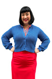 A smiling woman with black hair wears the Cissi och Selma cardigan Sigrid Hav by BUTIK KUBIK over a patterned blouse and red skirt, standing hands on hips against a white background.