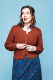 A woman with wavy brown hair wears the rust-colored Cissi och Selma cardigan Sigrid Kotte from BUTIK KUBIK over a blue dress with a red pattern made from ekologisk bomull, standing against a blue background and looking to the side with a surprised expression.