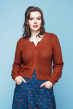 A woman with wavy brown hair is wearing the Sigrid Kotte cardigan from Cissi och Selma by BUTIK KUBIK in a rust color, layered over a blue dress with a red pattern. She stands against a light blue background, looking at the camera with her hands tucked into her dress pockets.