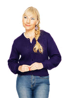 A woman with blonde hair in a side braid wears the Cissi och Selma cardigan Sigrid Lila by BUTIK KUBIK and blue jeans, standing against a white background and smiling softly while looking to the side.