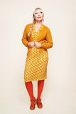 A woman with blonde braided hair, dressed in the Cissi och Selma cardigan Sigrid Senap from BUTIK KUBIK over a yellow patterned dress, red tights, brown shoes, and orange accessories, stands against a plain light background, looking up and smiling.