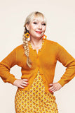 A smiling woman with blonde hair in a side braid wears the Sigrid Senap orange cardigan by Cissi och Selma from BUTIK KUBIK, a yellow patterned dress made from ekologisk bomull (organic cotton), and orange flower earrings. She poses with her hands on her hips against a plain background.