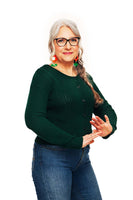 Older woman with long gray hair, glasses, and colorful earrings smiles while wearing the BUTIK KUBIK Cissi och Selma cardigan Sigrid Skog and jeans, standing against a plain white background with hands gesturing in front of her.