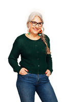 Older woman with long gray hair in a braid, wearing glasses, colorful earrings, blue jeans, and the BUTIK KUBIK Cissi och Selma cardigan Sigrid Skog, smiling with hands in her pockets against a white background.