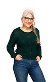 Older woman with long gray hair in a braid, wearing glasses, colorful earrings, blue jeans, and the BUTIK KUBIK Cissi och Selma cardigan Sigrid Skog, smiling with hands in her pockets against a white background.