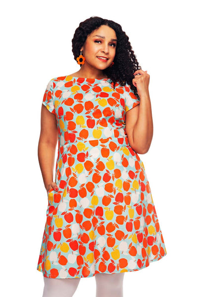 Woman wearing the BUTIK KUBIK Siv klänning Äppelriket, a 50s-inspired dress with a vibrant apple print in orange, red, and yellow. She has curly hair, orange earrings, white leggings, and is smiling while touching her hair.