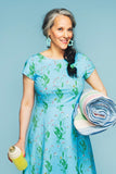 A smiling woman with gray-streaked dark hair in a side braid wears the Siv klänning Morgondagg by BUTIK KUBIK, holding a yellow bottle and a rolled-up striped blanket against a light blue background.