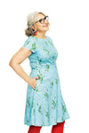 Smiling older woman with gray hair in a low bun, wearing glasses and the BUTIK KUBIK Siv klänning Morgondagg blue 1950s-inspired floral dress with hidden pockets, red earrings, and red leggings against a white background.