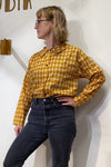 A person wearing glasses and the BUTIK KUBIK Kubik blouse vintage fabric—an oversized boxy-fit shirt with yellow and orange florals—paired with high-waisted black jeans, stands against a white wall looking to the side, one hand on their hip.