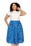A woman with curly hair wears a white short-sleeve top and the high-waisted, knee-length BUTIK KUBIK Cissi och Selma Solbritt skirt Ängsblom blå, featuring a white floral pattern, as she stands looking to the side with one hand in her hair.