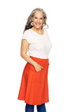 Smiling woman with long gray hair in a white top, blue leggings, and the orange Cissi och Selma Sonja skirt Apelsin by BUTIK KUBIK with pockets, stands with hands in her pockets against a white background.