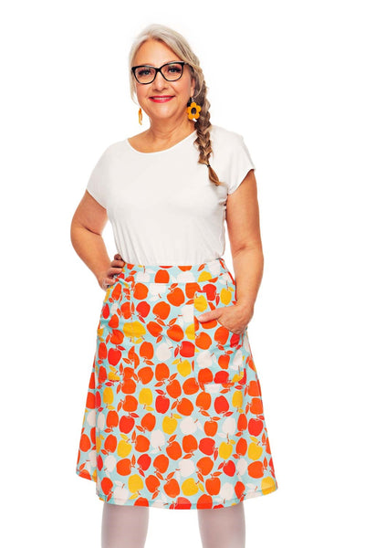Smiling older woman with gray braided hair, glasses, and a white top wears the colorful Cissi och Selma Sonja skirt Äppelriket from BUTIK KUBIK with pockets, standing with one hand in her pocket against a white background.