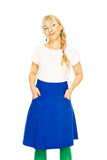 A blonde woman in a braid, wearing a white shirt and the BUTIK KUBIK Cissi och Selma Sonja skirt Blåsippa, green tights, and green earrings stands smiling with her hands in the skirt pockets against a white background.