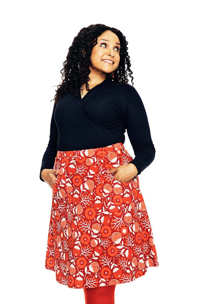 A woman with curly hair, smiling with hands in her pockets, wears a black top and the BUTIK KUBIK Cissi och Selma Sonja skirt Hassel röd—red with a white and orange floral pattern—against a white background.