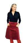 A smiling woman with long, gray-streaked hair wears a black top and the Cissi och Selma Sonja skirt corduroy rabarber from BUTIK KUBIK, featuring large pockets. She pairs it with red tights and stands against a white background.