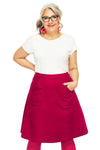 Smiling older woman with gray hair, glasses, and red earrings wears a white shirt and the BUTIK KUBIK Cissi och Selma Sonja skirt ros, a bright pink flared cotton skirt with pockets, standing against a white background with one hand in her pocket.