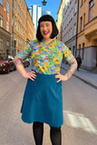 A smiling woman with black hair and visible tattoos stands in a city street under clear skies, wearing a colorful floral shirt, black tights, and the BUTIK KUBIK Cissi och Selma Sonja kjol Manchester Petrol. Tall buildings line the background.