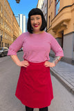 A woman with black hair, wearing a red and white striped top and the Cissi och Selma Sonja kjol Manchester Ros from BUTIK KUBIK, smiles on a city street lined with yellow and beige buildings.
